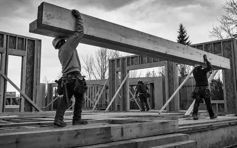 Builders FirstSource US12189T1043 Zimmermänner tragen schwere Holzbalken auf einer Wohnhaus-Baustelle im Schwarzweißfoto - Foto: THN