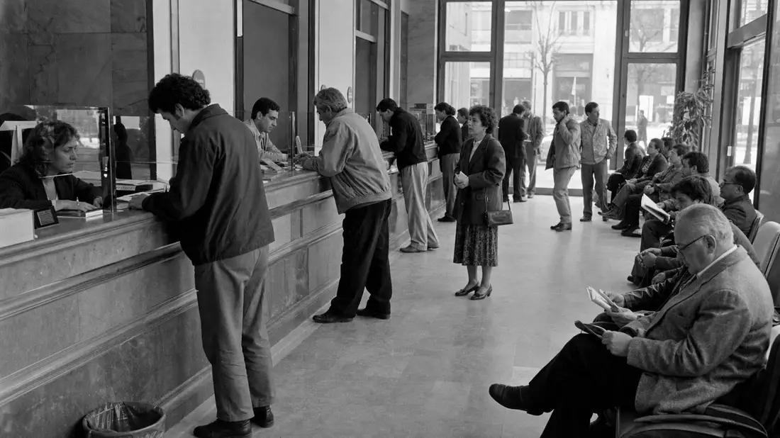 Schwarzweiß-Reportagefoto einer belebten Bankschalterhalle mit wartenden Kunden
