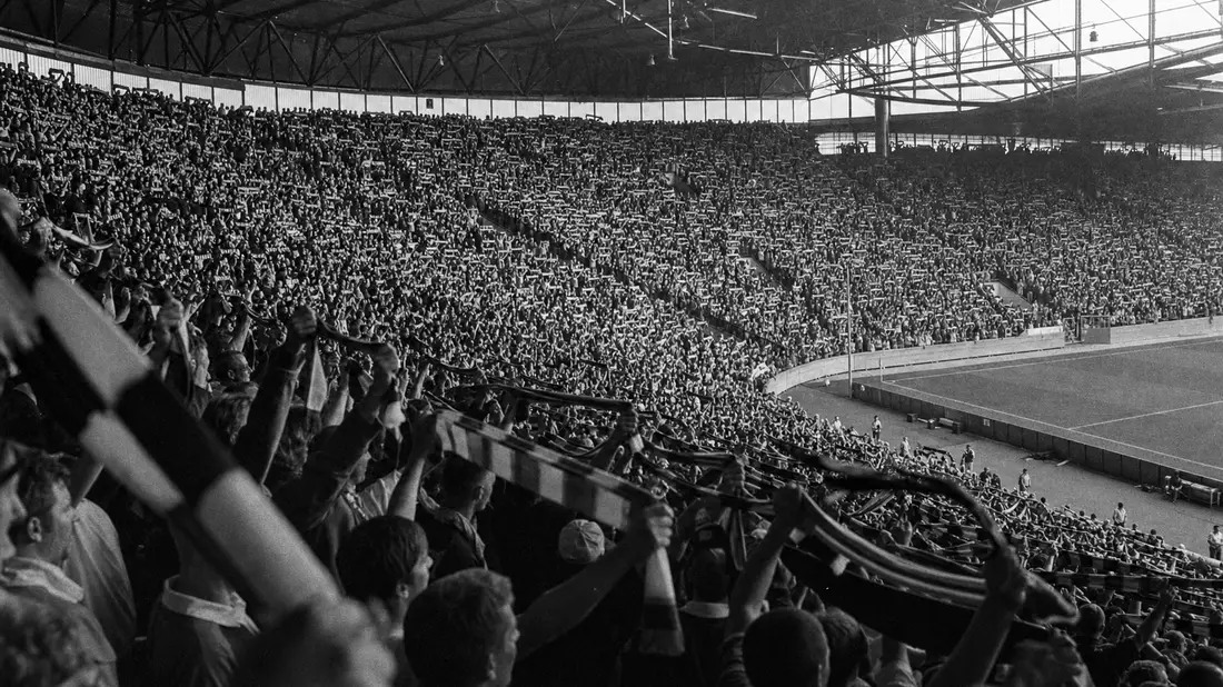 Schwarz-Weiß-Reportagefoto der vollbesetzten Südtribüne mit jubelnden Fans und Schals