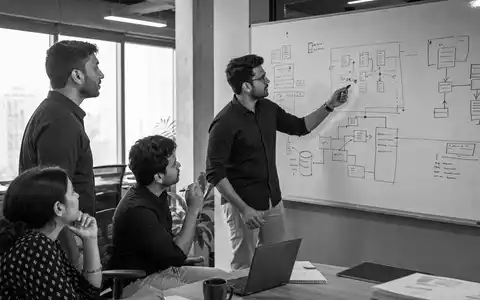 Schwarzweiß-Reportagefoto dokumentiert Teambesprechung bei Reply S.p.A., ISIN IT0005282865, vor Whiteboard mit Systemarchitektur-Diagrammen - Foto: THN