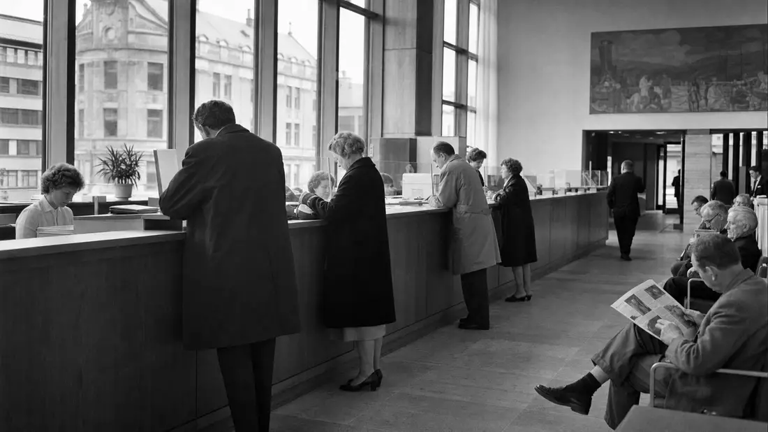 Dokumentarische Schwarzweißaufnahme einer Bankfiliale mit Kunden am Schalter