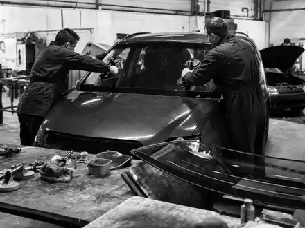 D'Ieteren Group BE0974259880 dokumentiert schwarzweiß mehrere Techniker bei sorgfältiger Windschutzscheiben-Reparatur in industrieller Werkstattumgebung mit Werkzeugen - Foto: THN