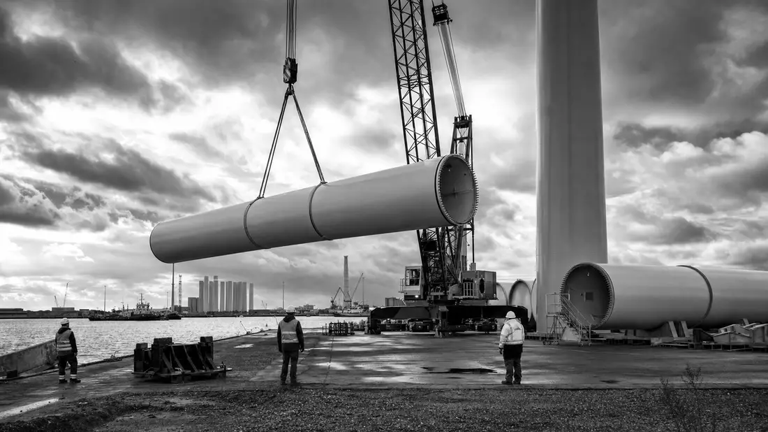 Schwarzweiß-Reportagefoto von Windturbinen-Bauteilen am Hafen mit Kran und Arbeitern