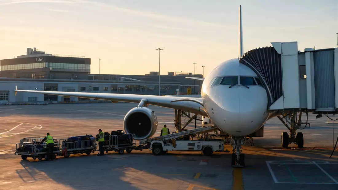 Weißes Kurzstreckenflugzeug am Flughafen Luton in warmem Morgenlicht, Gepäckwagen davor