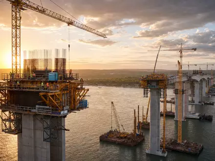 HOCHTIEF AG Brückenbau-Großprojekt DE0006070006 zeigt Kräne und Betonstützen bei Sonnenuntergang über dem Fluss - Foto: THN