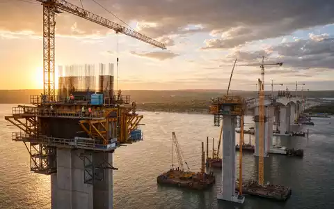 HOCHTIEF AG Brückenbau-Großprojekt DE0006070006 zeigt Kräne und Betonstützen bei Sonnenuntergang über dem Fluss - Foto: THN
