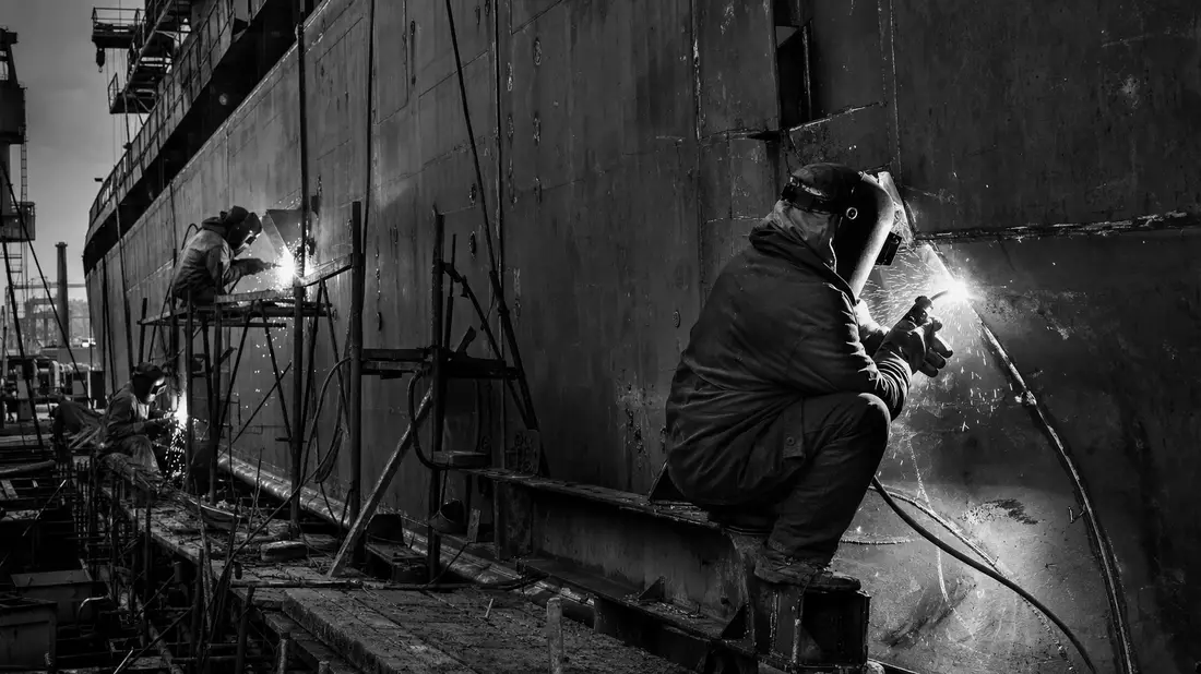Schwarzweiß-Reportage: Schweißer mit Funkenregen an massivem Stahlschiffsrumpf in der Werft