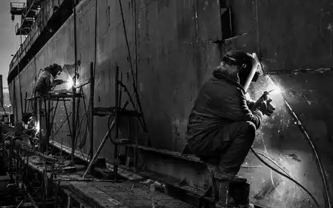 Huntington Ingalls Schweißer arbeiten in Schwarzweiß an einem massiven Schiffsrumpf in der Werft US4464131063 - Foto: THN