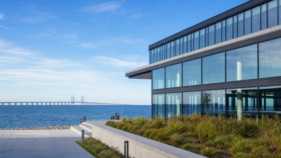 Modernes Glasbürogebäude am Wasser mit Öresundbrücke im Hintergrund, blauer Himmel