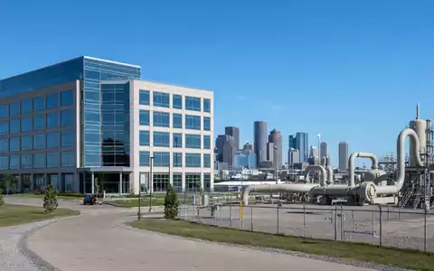 Loews Corporation US5404241031 zeigt fotorealistisch ein modernes Bürogebäude neben einer Erdgas-Pipelineanlage vor der Stadtskyline - Foto: THN