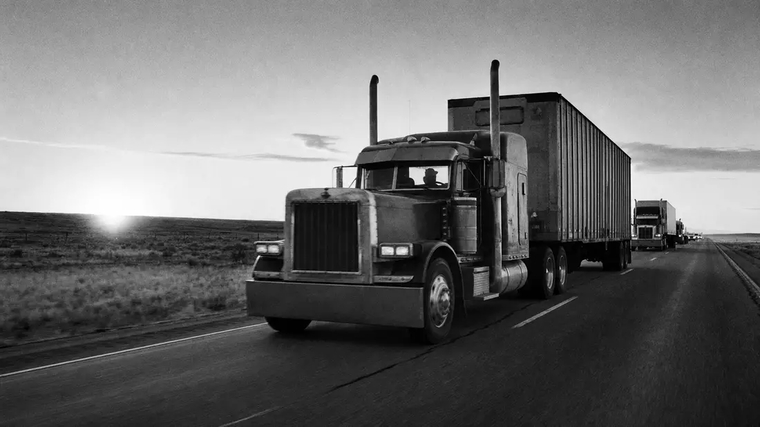Schwarz-Weiß-Reportagefoto eines LKW-Konvois auf einer Landstraße bei Sonnenaufgang
