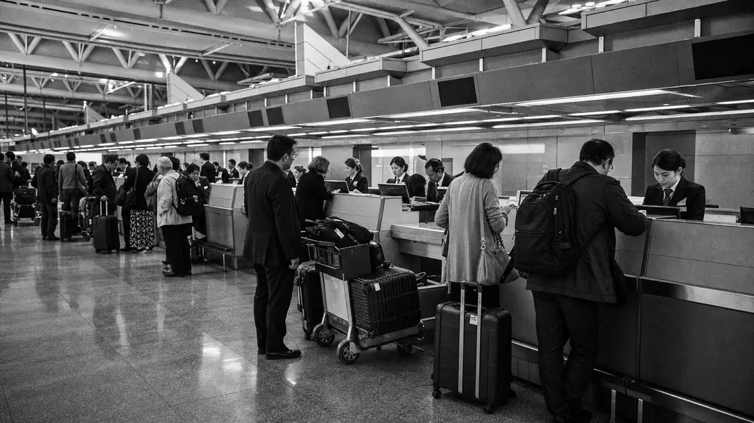 High-contrast black and white documentary photograph of busy airline check-in hall, uniformed agents at long counter row, passengers with luggage queuing