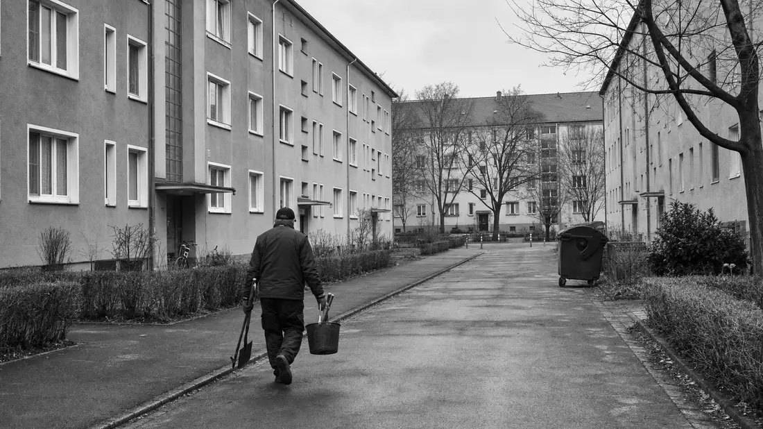 Schwarzweiß-Reportagefoto einer Wohnsiedlung mit Hausmeister auf der Straße