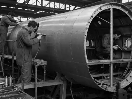 Dassault Aviation SA (FR0000121725): SchwarzweiĂ-Reportage dokumentiert Fabrikarbeiter beim prĂ€zisen Nieten einer metallischen Flugzeugrumpfsektion - Foto: THN