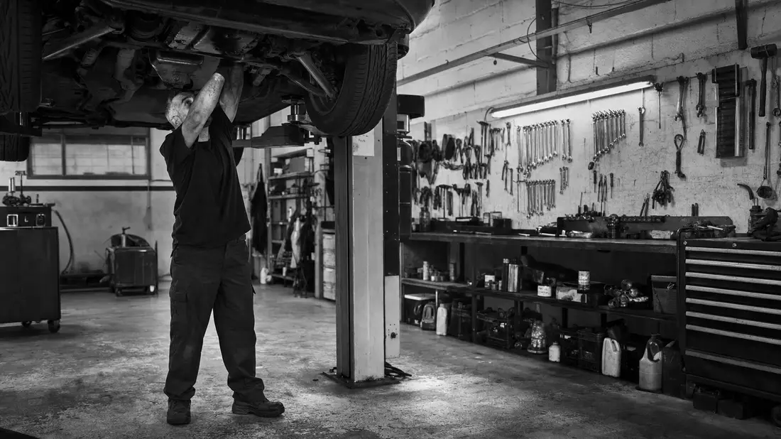 Schwarzweißfoto eines Mechanikers, der unter einem angehobenen Fahrzeug in der Werkstatt arbeitet