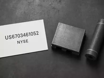 Nucor Corporation US6703461052 als Flatlay mit ISIN-Karte, Stahlbrammen-Miniatur und Grafitelektrode auf dunklem Untergrund - Foto: THN