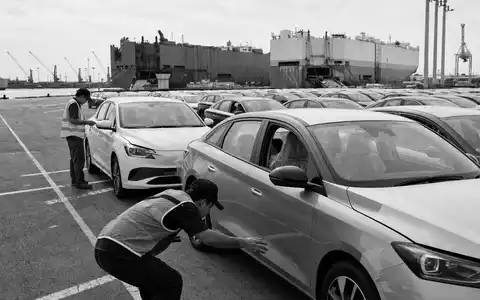 Inchcape plc GB00B61TVQ02 dokumentiert eine Schwarzweiß-Reportage-Aufnahme von Arbeitern bei sorgfältiger Fahrzeugkontrolle direkt am Hafen - Foto: THN