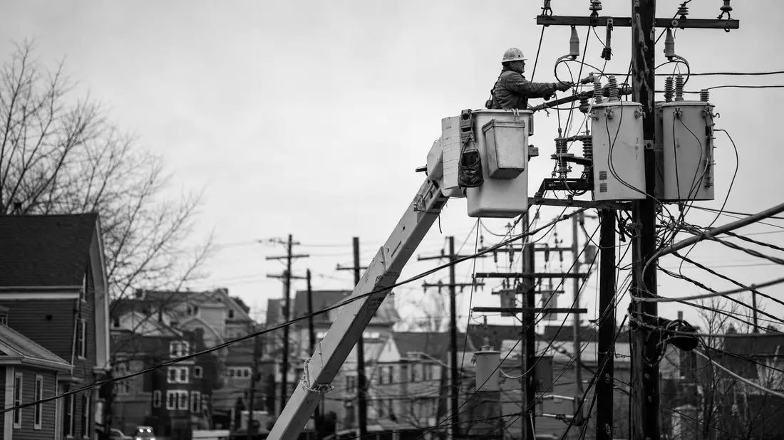 Schwarzweiß-Reportage: Elektriker im Arbeitseimer an Strommast in Vorstadtstraße Neuenglands