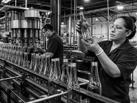 Diageo plc GB0002374006 dokumentiert schwarzweiß eine Abfüllanlage, in der Arbeiter unbeschriftete Glasflaschen auf Qualität prüfen - Foto: THN