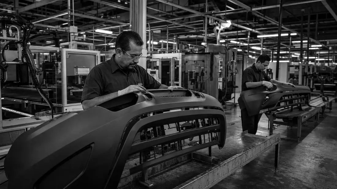 Schwarzweiß-Dokumentarfoto von Arbeitern an Kunststoff-Stoßfängern in Fabrikhalle