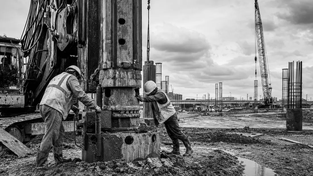 Schwarzweiß-Dokumentarfoto von Bauarbeitern an schwerer Bohrmaschine auf Großbaustelle