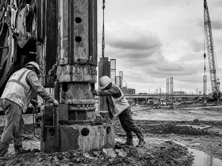 Schwarzweiße Reportage-Aufnahme dokumentiert Bauarbeiter an Fundamentbohrer, symbolisch für Keller Group plc, ISIN GB0034293025 - Foto: THN