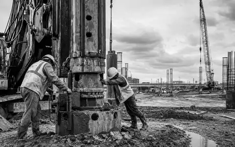 Schwarzweiße Reportage-Aufnahme dokumentiert Bauarbeiter an Fundamentbohrer, symbolisch für Keller Group plc, ISIN GB0034293025 - Foto: THN