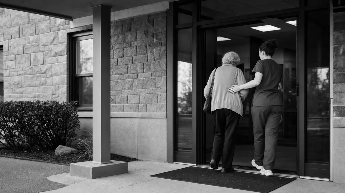 Schwarzweiß-Reportagefoto einer Pflegekraft, die eine ältere Patientin zum Eingang begleitet
