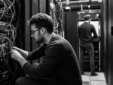 Computacenter plc GB00BV9FP302 dargestellt als dokumentarische Schwarzweiß-Reportage von zwei IT-Technikern im engen Serverraum - Foto: THN