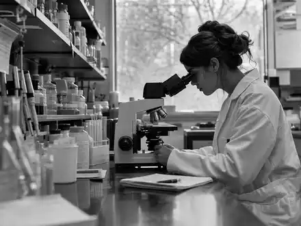 Coloplast A/S (DK0060448595) inspiriert diese dokumentarische Schwarzweiß-Aufnahme eines konzentrierten Forschungslabors mit Wissenschaftlerin am Mikroskop - Foto: THN