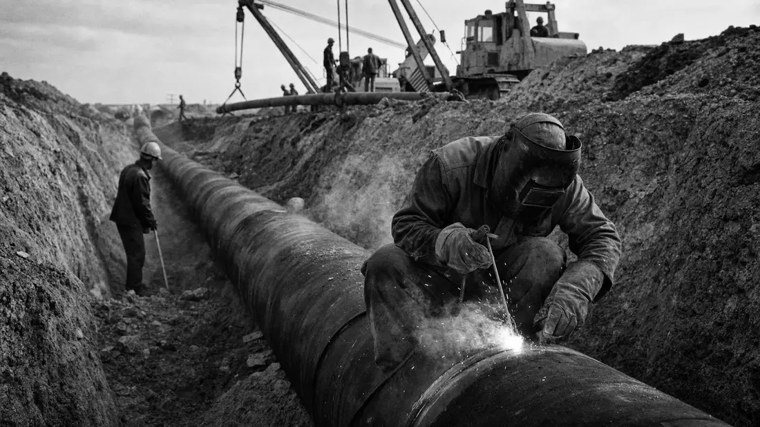 Schwarzweiß-Reportagefoto eines Schweißers bei der Pipeline-Verlegung im Erdgraben