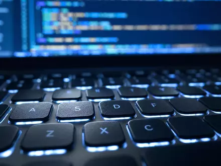 Paylocity Holding US70436Y1038 zeigt Makroaufnahme einer beleuchteten Laptop-Tastatur mit unscharfem Quellcode im Hintergrund - Foto: THN