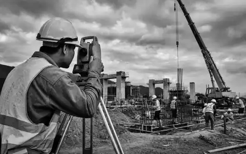 Trimble Inc. (US8962391058) dokumentiert Baubranche: Schwarzweiß-Reportagefoto eines Ingenieurs mit Tachymeter auf Infrastrukturbaustelle - Foto: THN