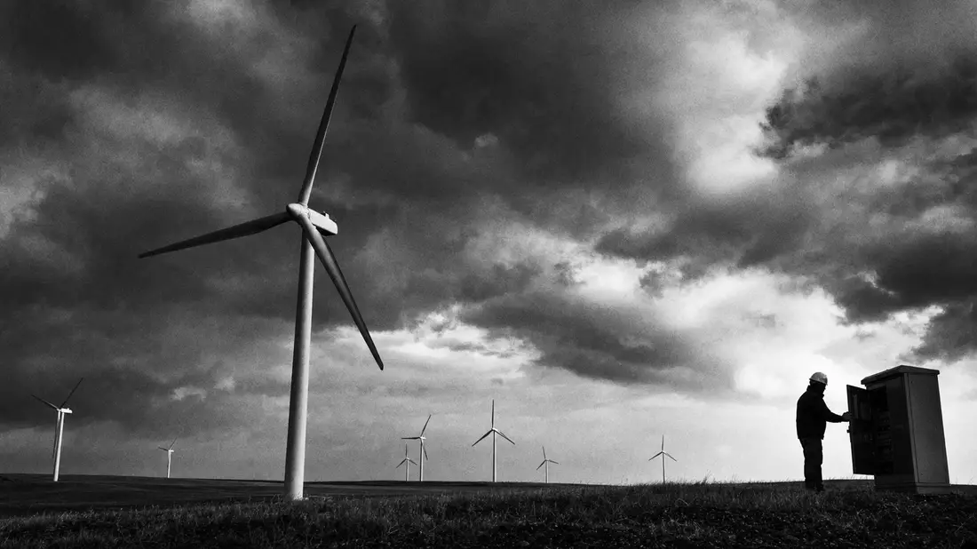Schwarz-weißes Reportagebild von Windturbinen mit Techniker unter stürmischem Himmel