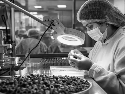 Schwarzweiß-Reportage zeigt Qualitätskontrolle bei West Pharmaceutical (US9523901012), Prüfung von Gummi-Verschlüssen unter Lupenlampe - Foto: THN