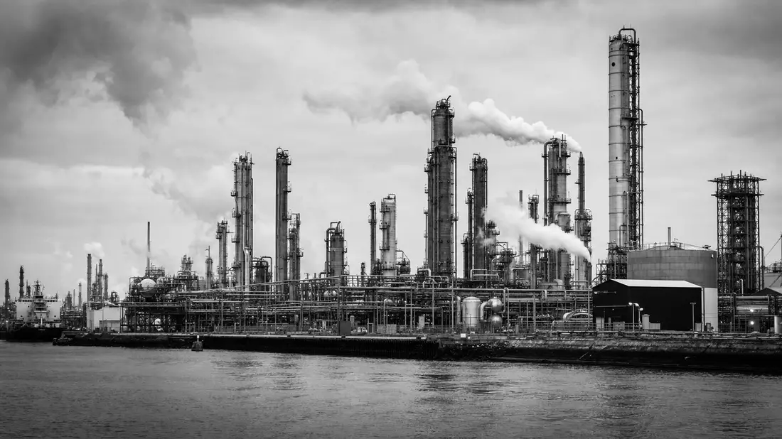 Schwarzweiß-Dokumentarfoto einer großen Chemieanlage am Flussufer mit Rohrleitungen und Türmen