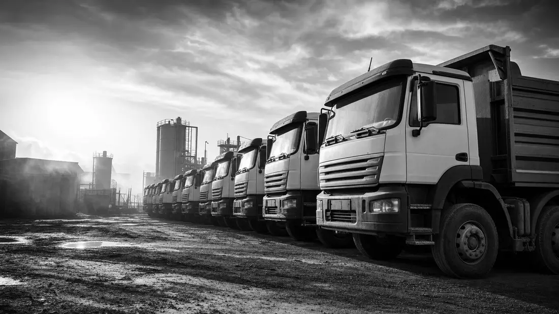 Schwarz-Weiß-Reportagefoto einer Flotte schwerer LKW auf industriellem Außengelände bei Gegenlicht