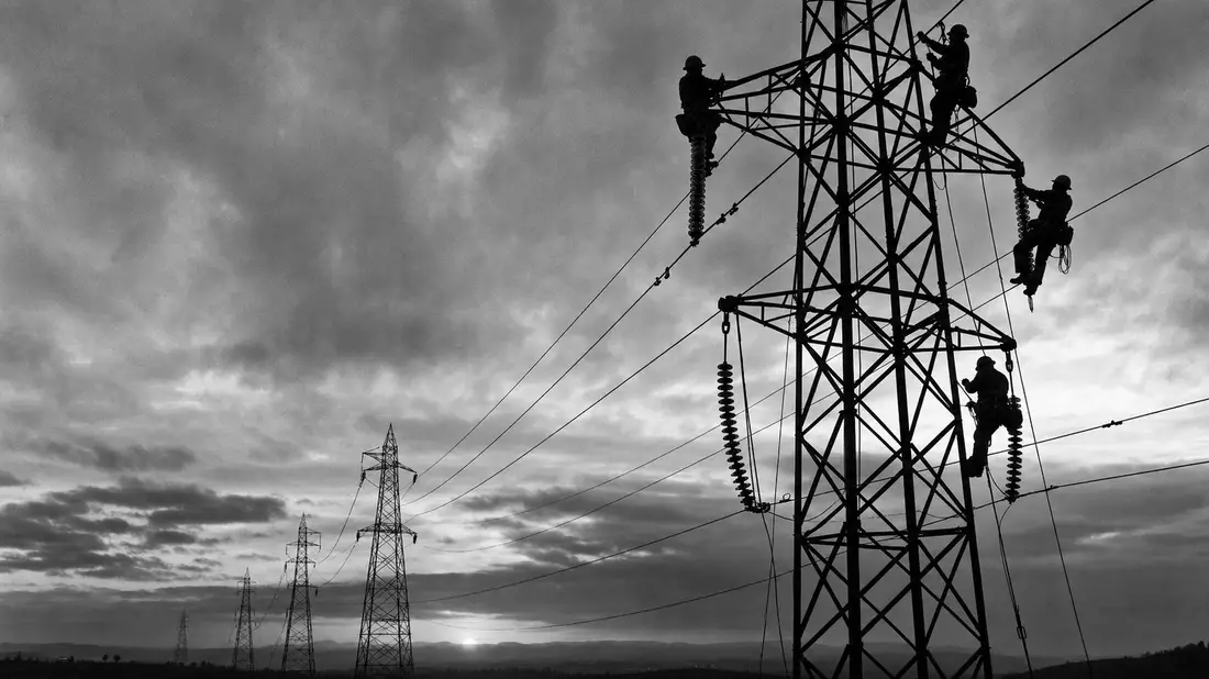 Schwarzweiß-Reportage: Monteure klettern an Hochspannungsmasten bei bewölktem Himmel