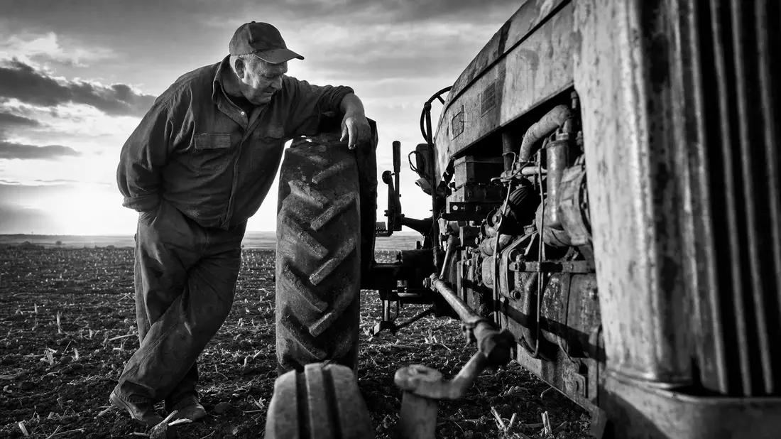 Schwarzweiß-Dokumentarfoto eines Landwirts neben schwerem Traktor auf Feld
