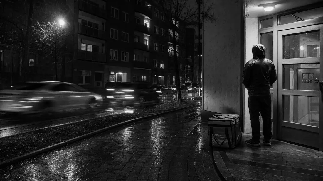 Schwarz-Weiß-Reportage: Lieferant wartet nachts im Regen vor Wohnhaus