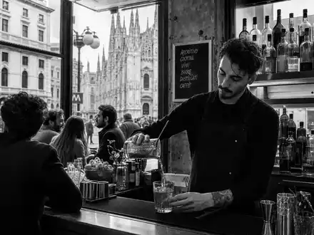 Davide Campari-Milano N.V. (NL0015435975) dokumentiert schwarzweiße Reportage-Aufnahme einer belebten Bar-Szene mit Mailänder Dom im Hintergrund - Foto: THN