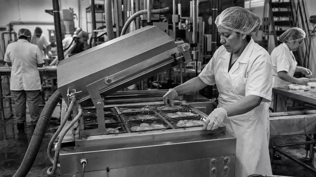 Schwarzweiß-Reportagefoto einer Arbeiterin an Vakuumversiegelungsmaschine in Lebensmittelfabrik