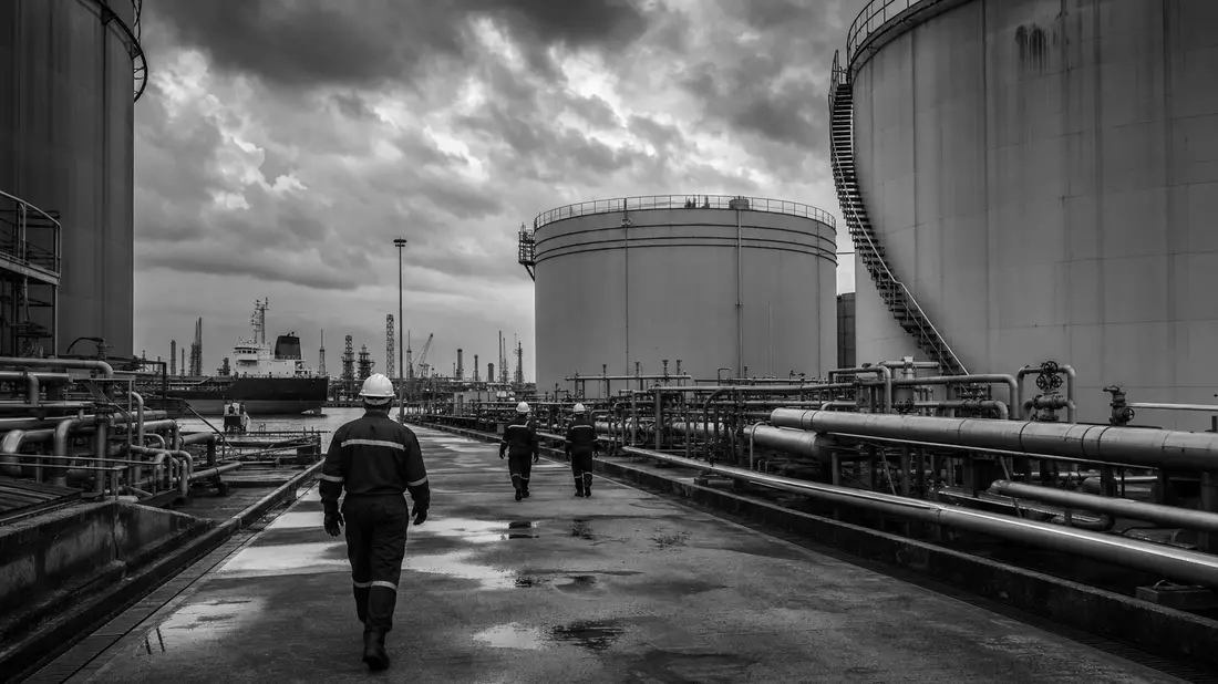 Schwarzweiß-Reportagefoto von Arbeitern zwischen Tanklager-Anlagen im Hafen