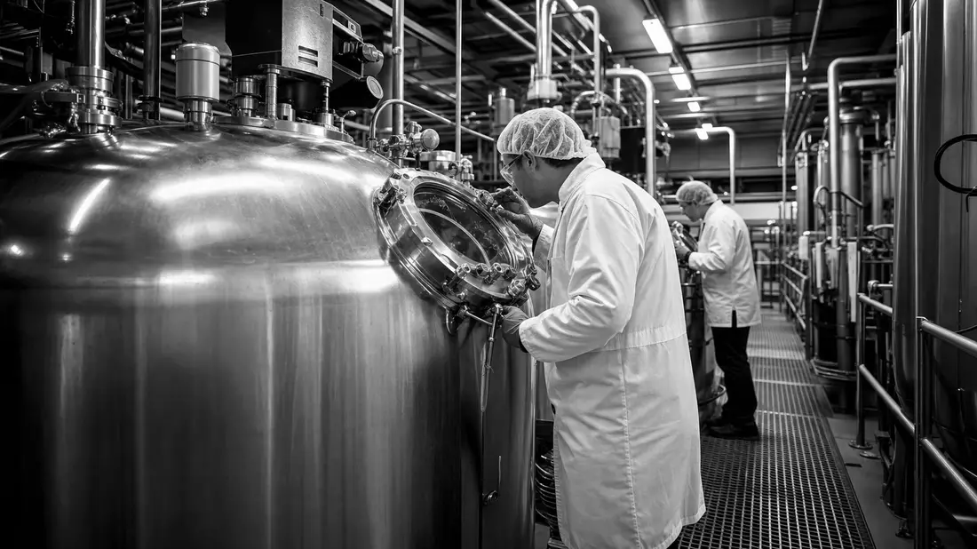 Schwarzweiß-Reportagefoto von Arbeitern an Fermentationstanks in Schutzkleidung