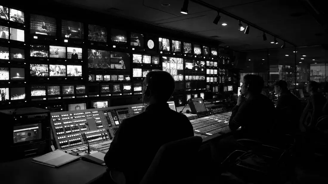 Schwarzweiß-Aufnahme eines Regieraums mit Monitorwand und Technikern bei der Arbeit