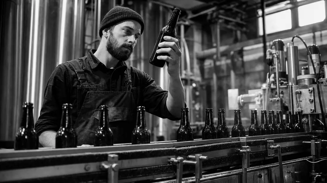 Schwarz-Weiß-Reportagefoto eines Arbeiters bei der Flaschenkontrolle in einer Brauerei