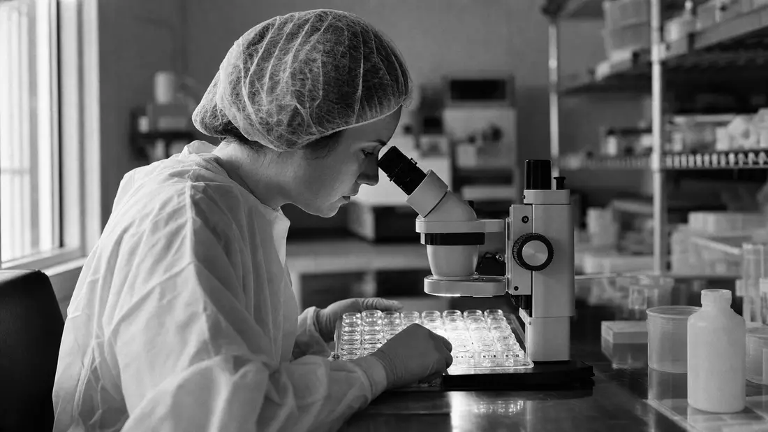 Schwarzweiß-Reportagefoto einer Wissenschaftlerin, die Kontaktlinsenformen unter dem Mikroskop prüft