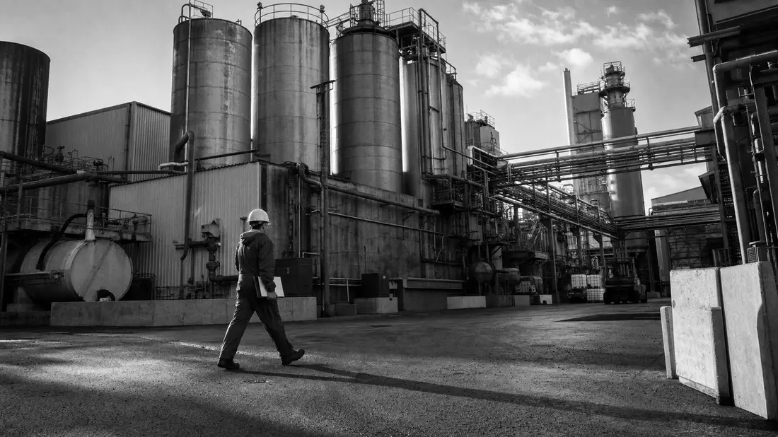 Schwarzweißfoto eines Arbeiters vor großen Industriesilos einer Chemiefabrik