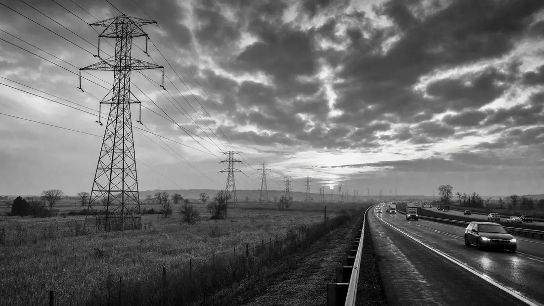 Dokumentarische Schwarzweißaufnahme von Strommasten entlang einer Autobahn bei Sonnenaufgang