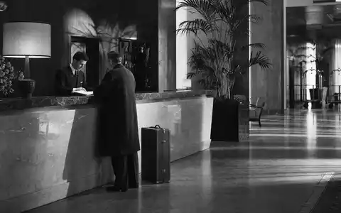 Host Hotels & Resorts US44107P1049 – dokumentarische Schwarzweiß-Reportage mit Gast und Concierge an klassischem Hotelempfang - Foto: THN